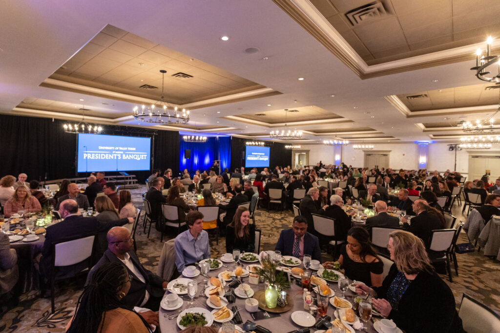 The banquet hall buzzes as elegantly dressed guests gather around tables, dining under chandeliers with Presidents Banquet displayed.