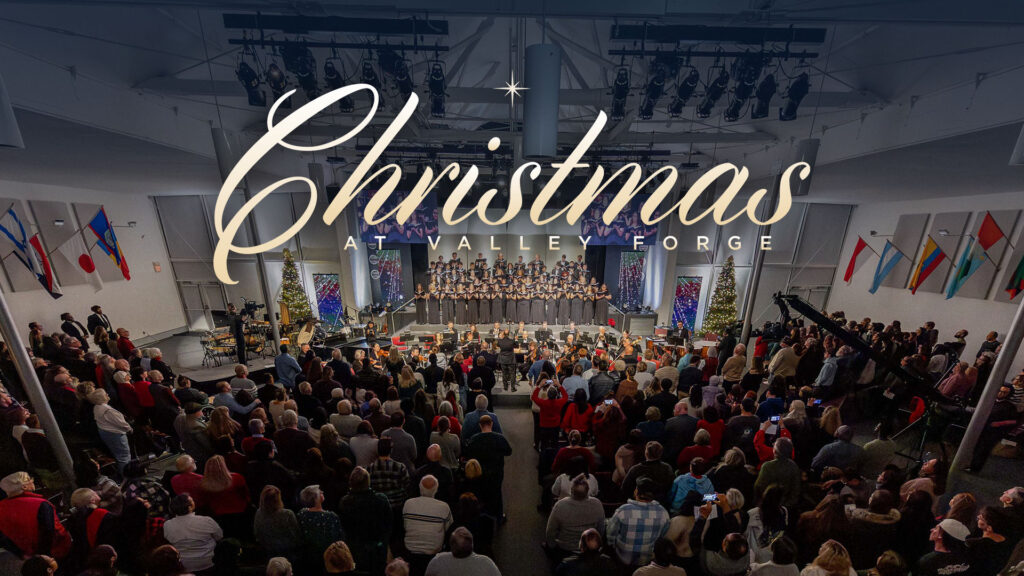 A festive indoor event features a choir on stage. Christmas at Valley Forge lights up the scene as the audience gathers.