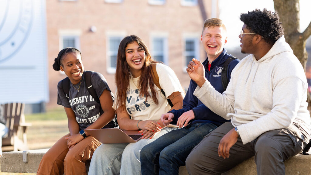 Four students from Right College sit on a low wall outdoors, smiling and laughing with a laptop. A building adds to the relaxed vibe.