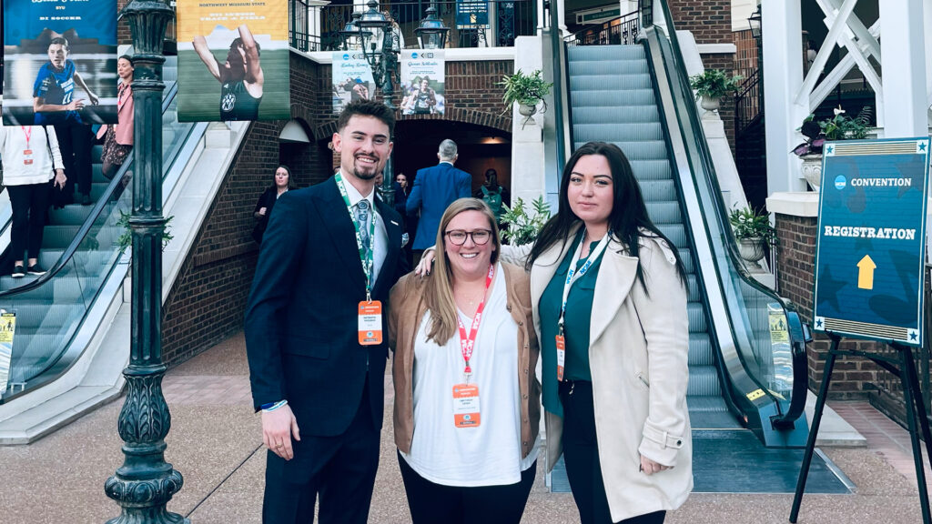 Three smiling people in business attire pose by escalators for the NCAA Convention; a registration sign is visible nearby.