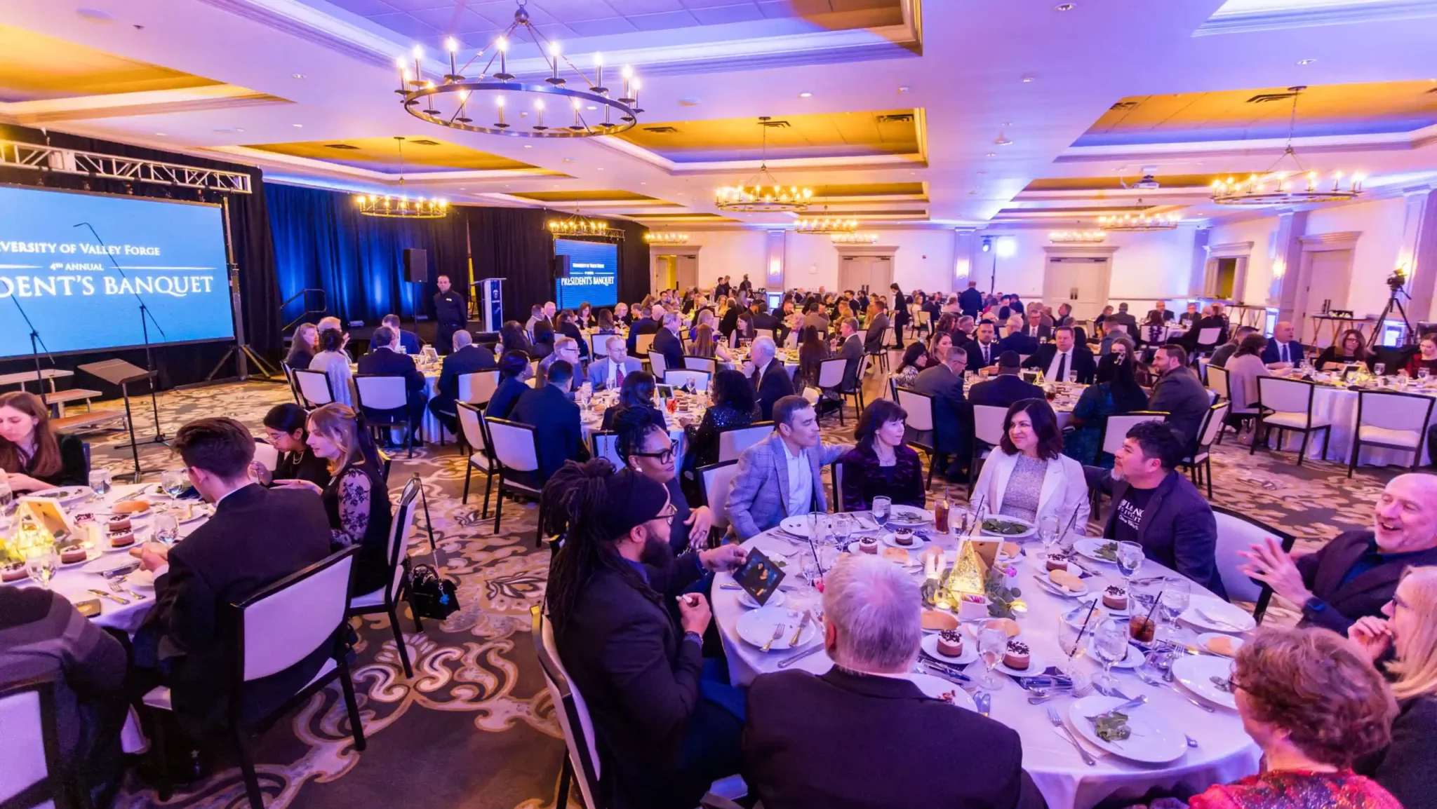 In a warmly lit banquet hall, the President's Banquet unfolds at round tables. The Presidents Banquet screen glows as guests chat.