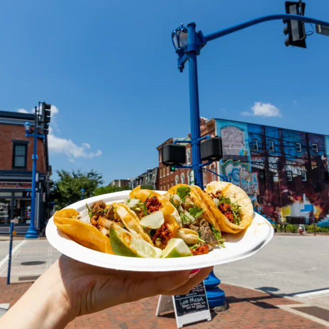 A hand holds tacos with cilantro, onions, and lime. Behind is a lively street scene with a mural and blue streetlights under the sky.