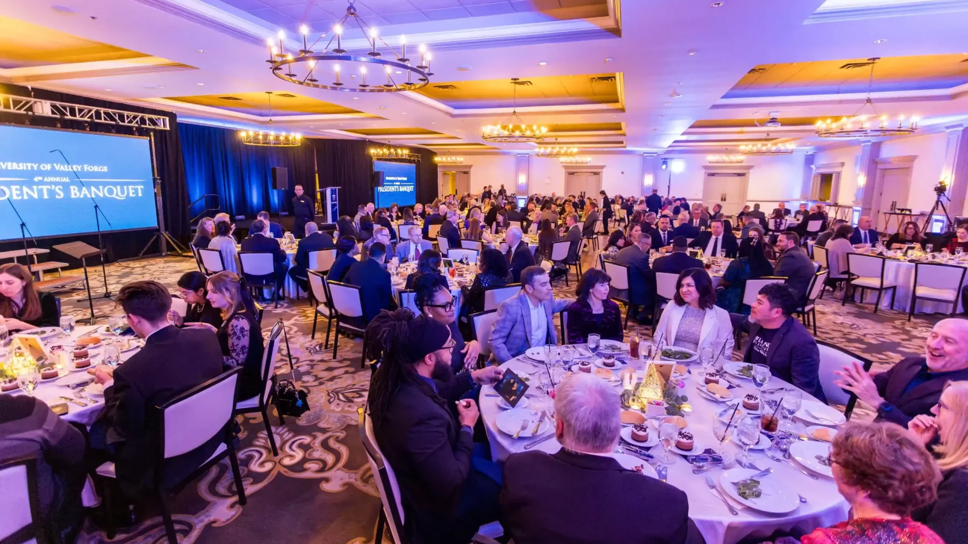 In a warmly lit banquet hall, the President's Banquet unfolds at round tables. The Presidents Banquet screen glows as guests chat.